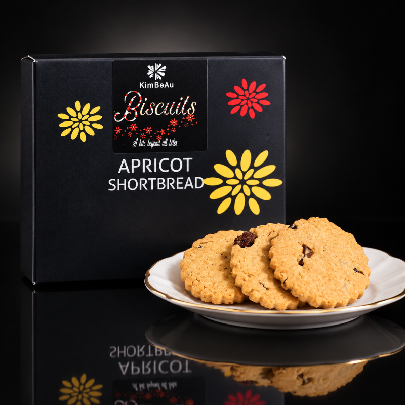 A black KimBeAu Biscuits box labeled 'Apricot Shortbread' stands behind a white plate holding three golden-brown shortbread biscuits with visible dried fruit pieces, on a glossy black reflective surface.
