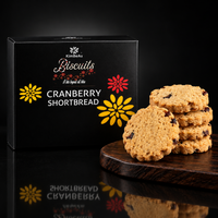 KimBeAu Cranberry Shortbread Biscuits, featuring a black packaging box with yellow and red flower graphics alongside several golden-brown shortbread cookies studded with cranberries on a dark wooden board against a gifted black box background. 