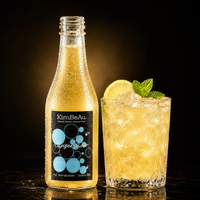 A bottle of KimBeAu Ginger Beer next to a glass of the golden beverage filled with ice, garnished with a lime slice and mint leaf, on a dark reflective surface against a black background.