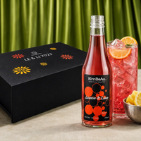 KimBeAu Lemon & Lime sparkling drink in a clear glass bottle with a black label, shown next to a pink drink in a glass with orange slices and lemon wheels, with a black LE & LI 7923 gift box in the background against a green curtain backdrop.