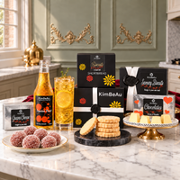 A styled kitchen countertop display featuring KimBeAu baked goods and a beverage. Visible items include boxes of shortbread, savoury biscuits, guava cheese, and chocolates, a bottle of passion fruit-flavoured non alcholic  drink, a glass of passion fruit drink with a lemon slice, shortbread biscuit on a marble board, and a plate of sugar-coated guava balls.