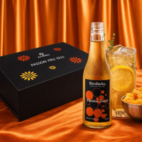 A bottle of KimBeAu Passion Fruit non-alcoholic beverage stands in front of a black gift box labeled PASSON FRUIT, with a glass of iced refreshing passion fruit drink and a bowl of fruit on an orange satin backdrop.