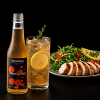 A bottle of KimBeAu Passion Fruit soda next to a glass of the golden refreshing drink with a lemon slice, set against a dark background with a plate of grilled chicken and salad.