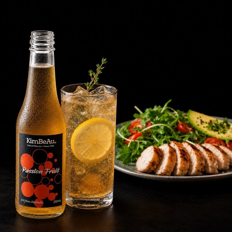 A bottle of KimBeAu Passion Fruit soda next to a glass of the golden refreshing drink with a lemon slice, set against a dark background with a plate of grilled chicken and salad.