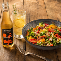 A styled tabletop scene with a bowl of mixed salad, a glass of lemonade with a lemon slice, and a bottle of KimBeau Passion Fruit Drink on a wooden table with a fork.