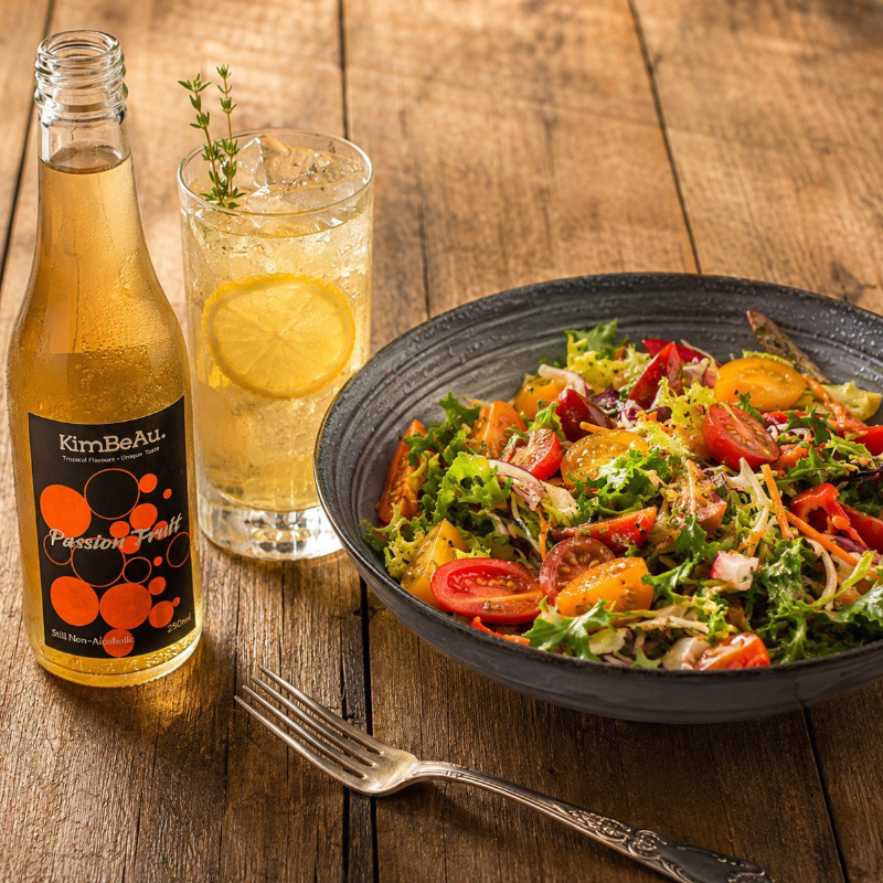 A styled tabletop scene with a bowl of mixed salad, a glass of lemonade with a lemon slice, and a bottle of KimBeau Passion Fruit Drink on a wooden table with a fork.