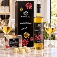 KimBeAu Passion F Blanc wine gift set displayed on a marble countertop with two filled wine glasses, a chocolate box, and several white chocolates. The set includes a black gift box with colorful floral graphics, a bottle of white wine, and a matching chocolate gift box.
