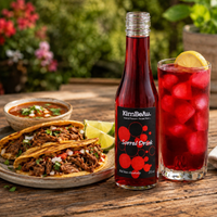 A bottle of KimBeAu Sorrel Drink stands on a wooden table beside a glass of the same red drink with ice and a lemon wedge, with authentic beef birria tacos and a bowl of salsa in the background.