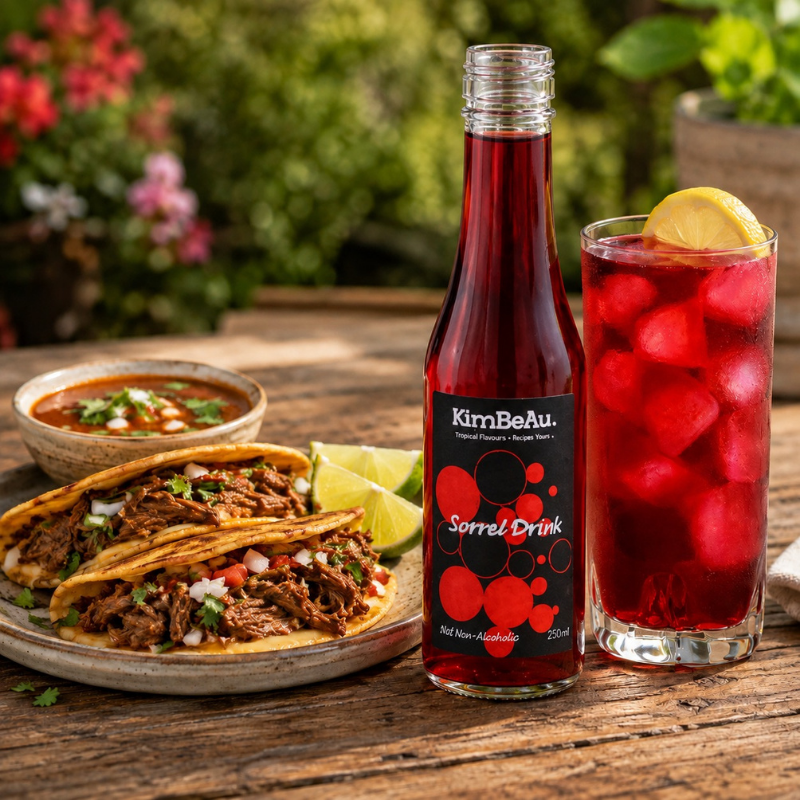A bottle of KimBeAu Sorrel Drink stands on a wooden table beside a glass of the same red drink with ice and a lemon wedge, with authentic beef birria tacos and a bowl of salsa in the background.