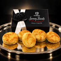 Five golden-brown savoury biscuits arranged on a reflective silver tray in front of a black gift box labeled KimBeAu Savoury Cheese Biscuits with a black rose and white ribbon, against a dark background.