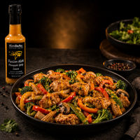 A bottle of KimBeAut Fusion Kick Pineapple Spice Sauce stands beside a large black plate of cooked chicken with broccoli, red bell peppers, mushrooms, and corn on a dark tabletop. A salad bowl and small dish of peppercorns appear in the background.